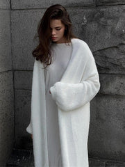 Woman wearing a long white fuzzy knit cardigan and a striped dress, standing against a stone wall.