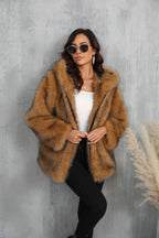 Woman wearing a brown faux fur hooded jacket with a white top and black pants, leaning against a gray wall.