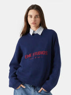 front view of a woman wearing a navy blue crew neck sweater with red "EME STUDIOS" embroidery