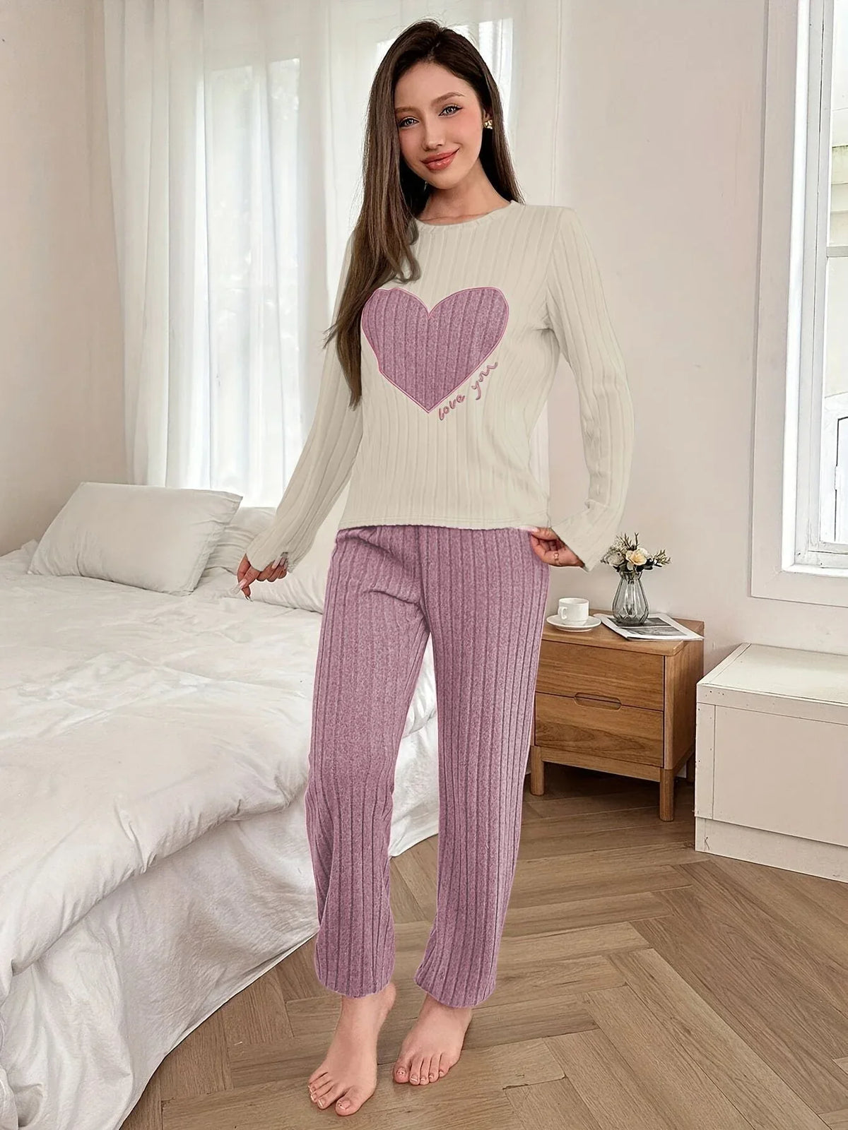 Woman standing in a bedroom wearing a cream long-sleeve pajama top with a pink heart graphic and "love you" text, and matching ribbed purple pajama pants. Main image