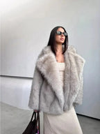 woman in a light grey faux fur coat and cream dress holding a brown bag