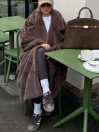 A woman enjoys a coffee at an outdoor cafe, enveloped in a luxurious brown faux fur coat.