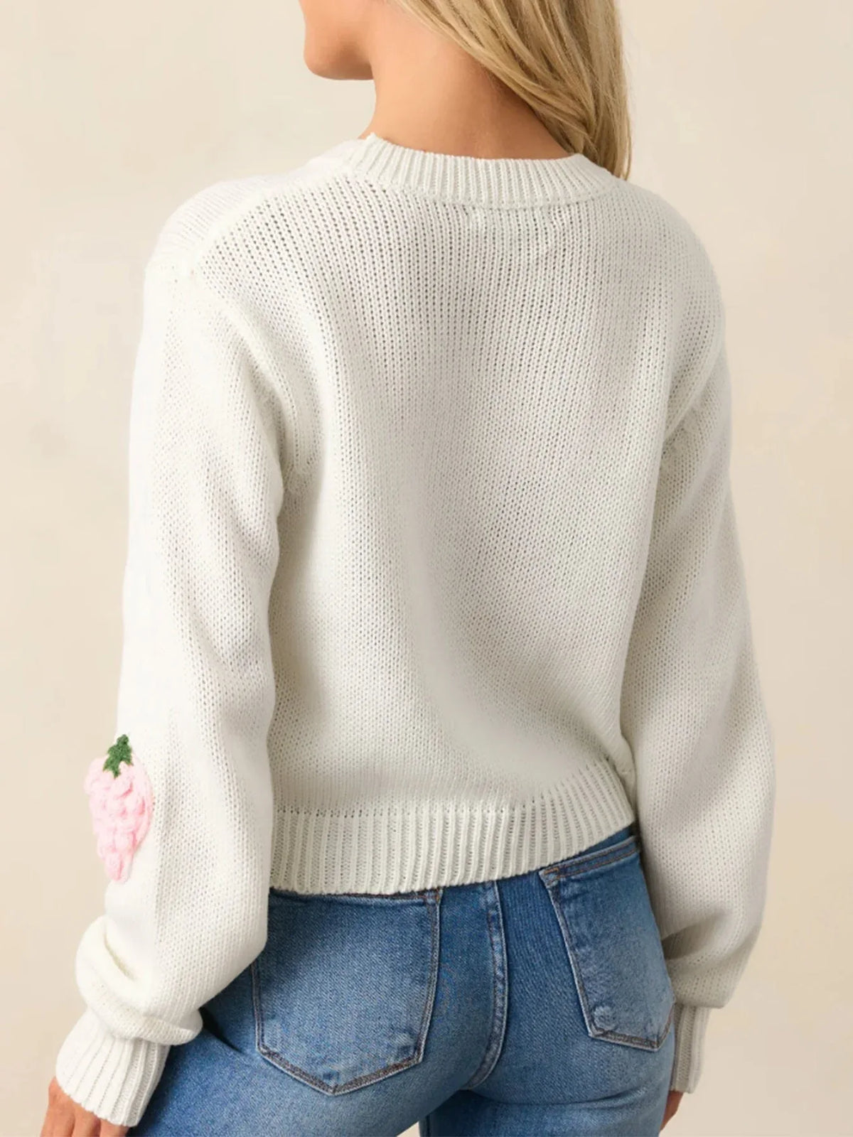 Back view of a white cropped knit sweater with a single pink strawberry applique on the sleeve, worn with blue denim jeans.