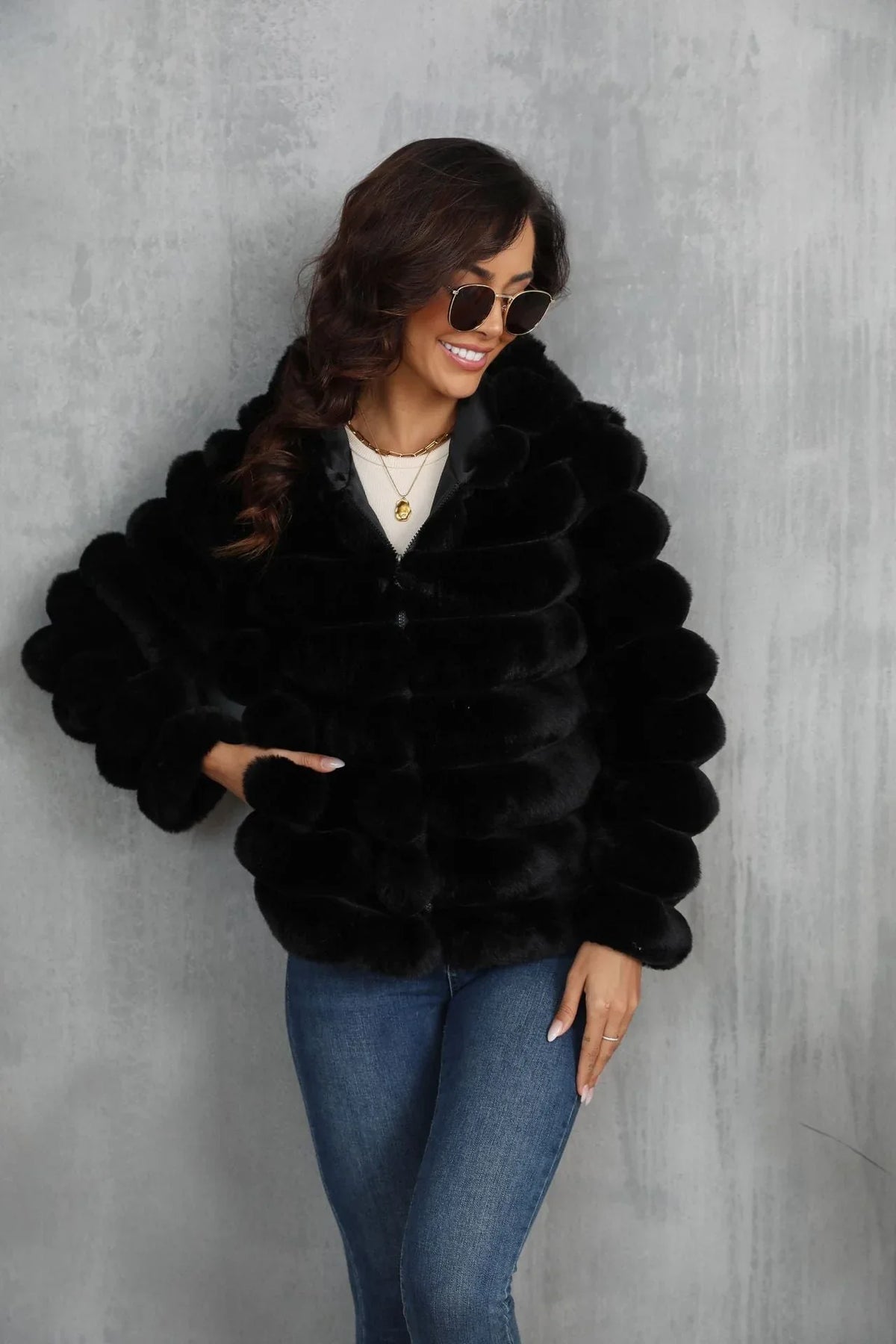 Woman wearing a black faux fur hooded jacket over a white crop top and blue jeans, with her hand in the pocket.