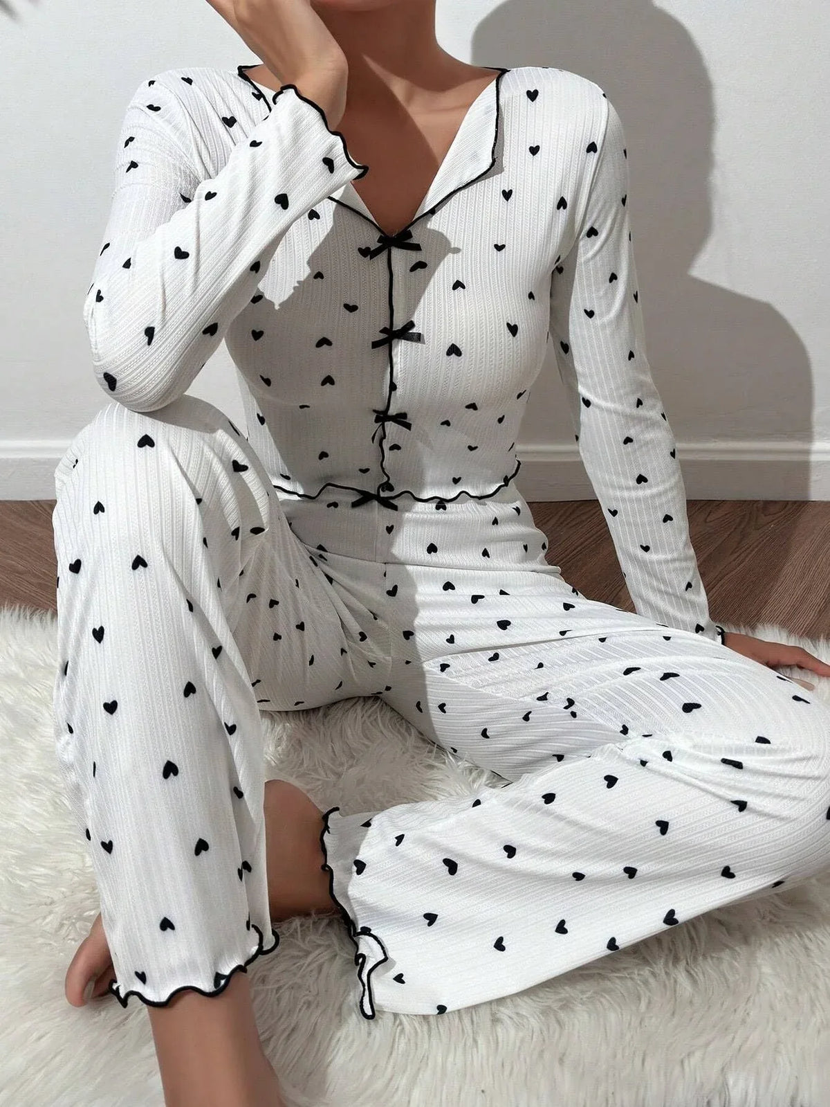 Woman wearing a white long-sleeve pajama set with a black heart print, sitting on a white rug.