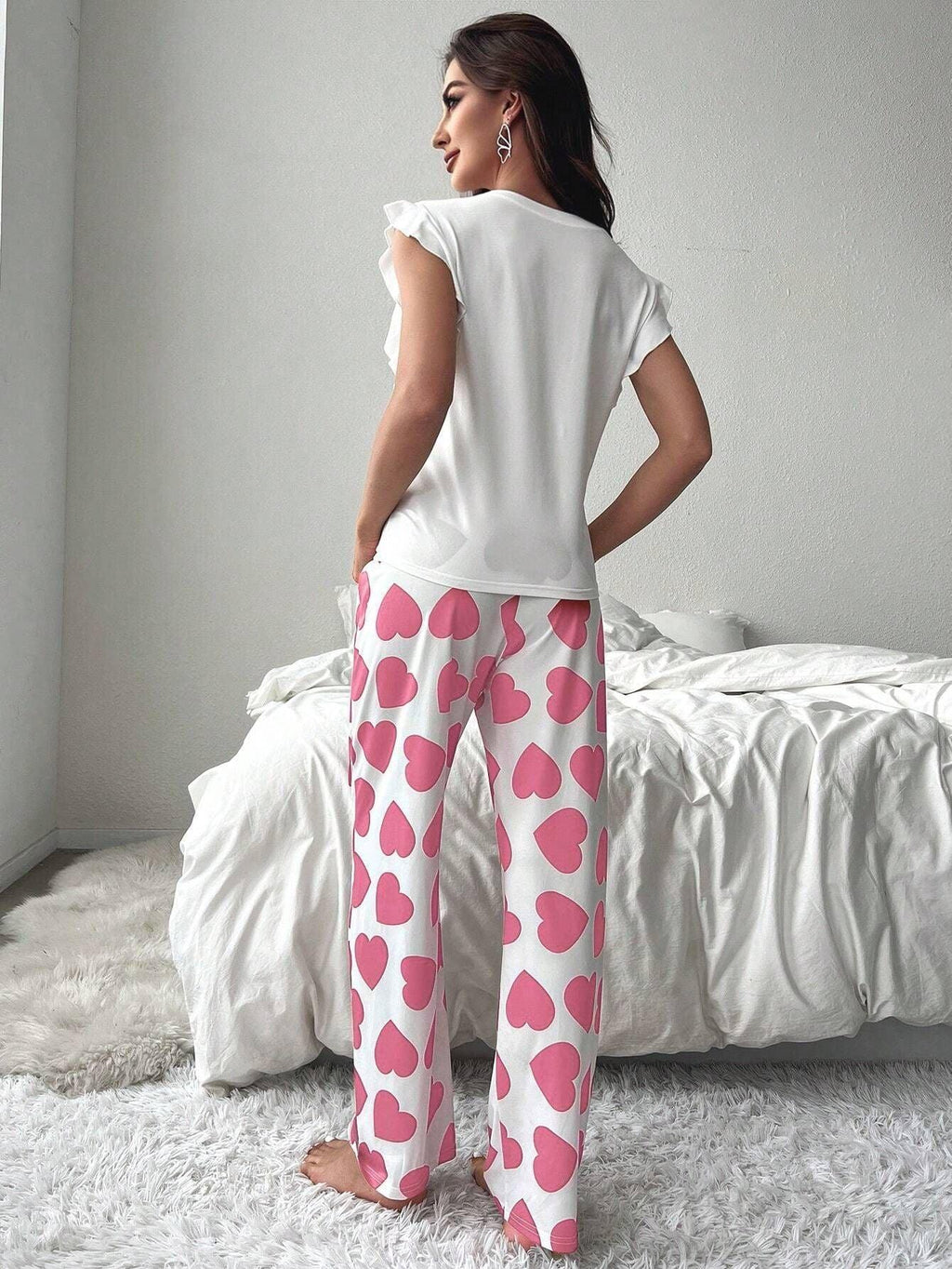 Back view of a woman standing next to a bed wearing white pajamas with pink heart print pants and a white top with a pink ribbon graphic.