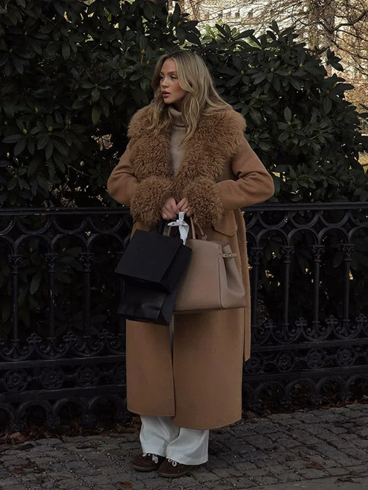 Woman in a long camel coat with faux fur trim, holding a black and a tan shopping bag.
