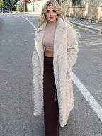 Woman wearing a cream faux fur long coat with a chevron pattern, tan crop top, and brown maxi skirt, standing on a street.