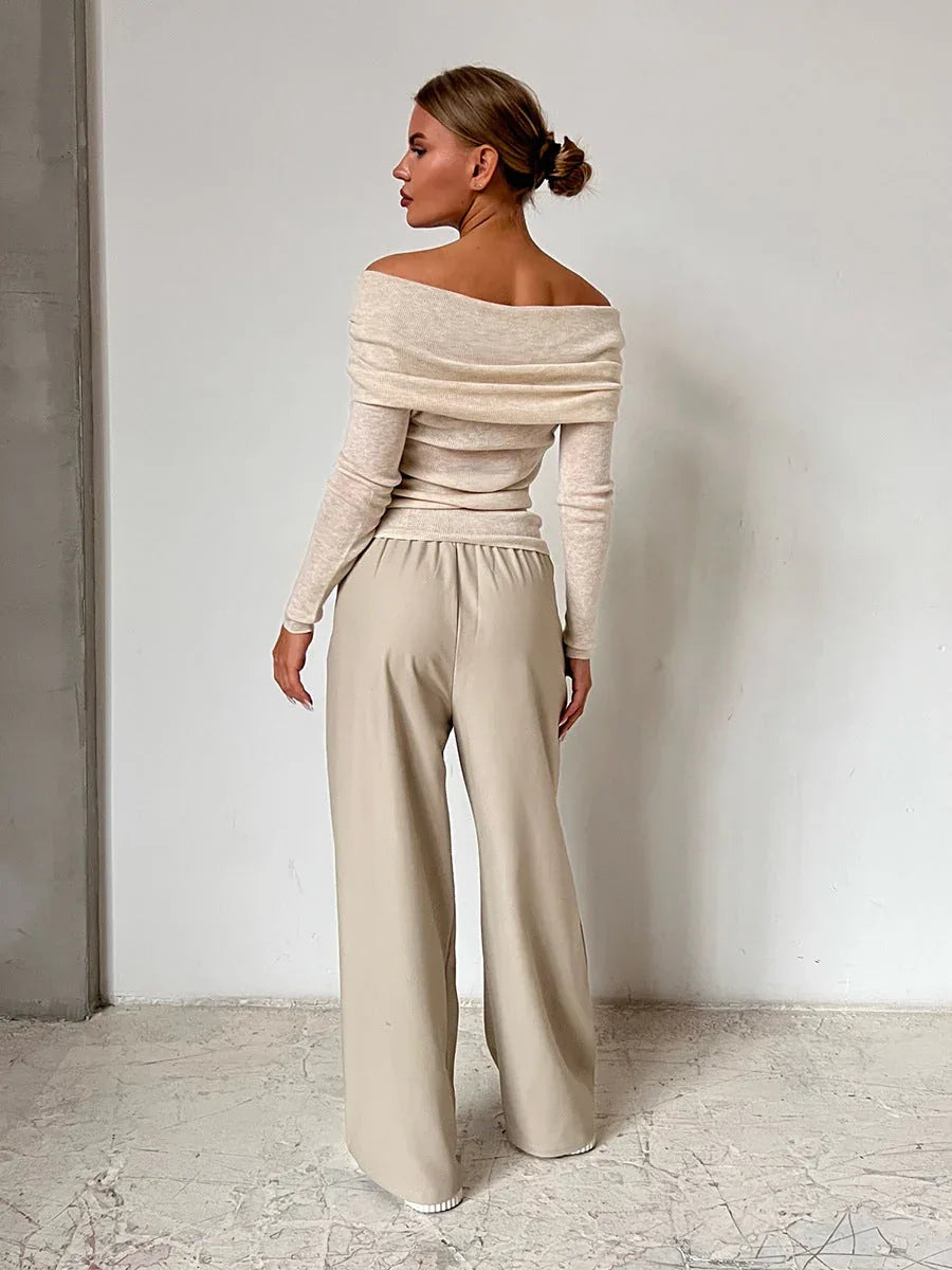 Back view of a woman wearing a cream off-the-shoulder ribbed knit sweater with long sleeves and beige wide-leg pants, standing against a white wall. Secondary image