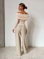 Back view of a woman wearing a cream off-the-shoulder ribbed knit sweater with long sleeves and beige wide-leg pants, standing against a white wall.