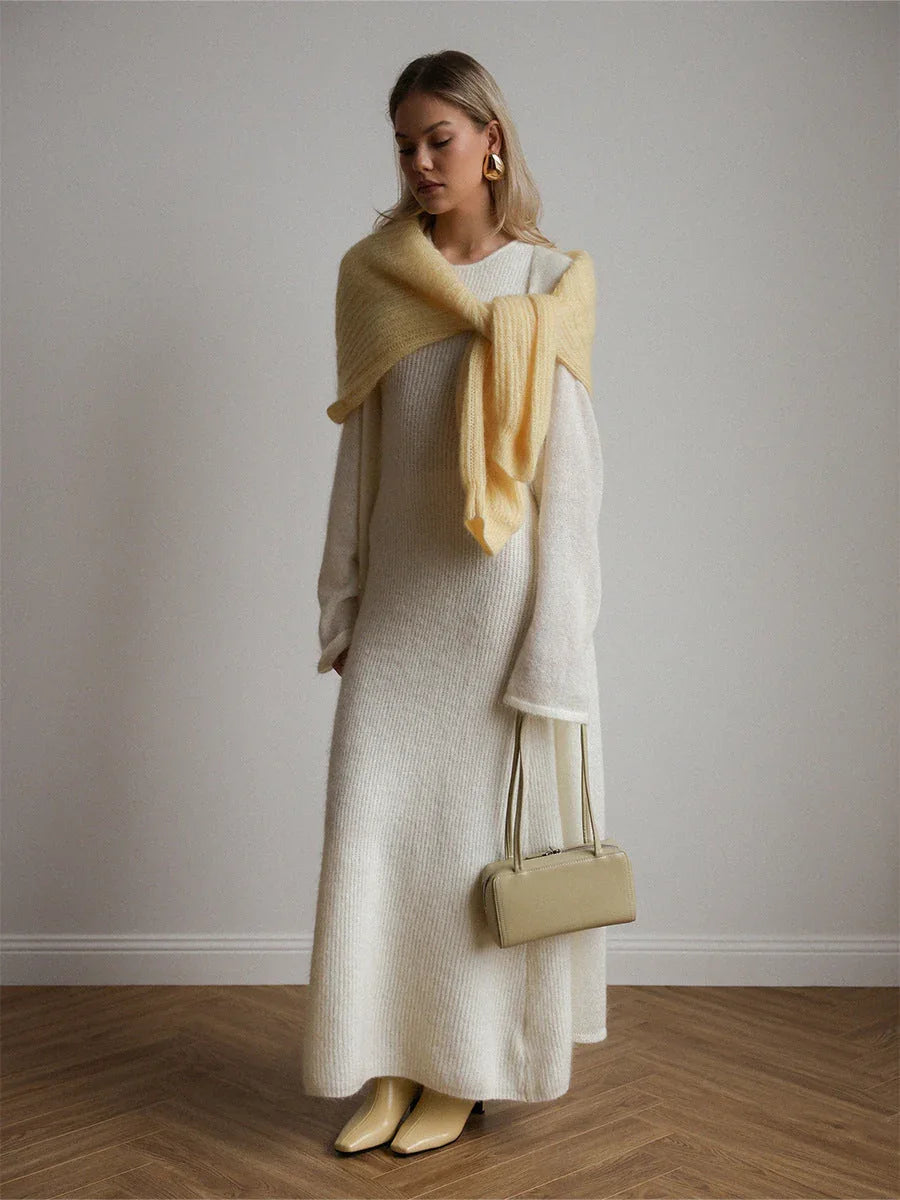 Woman wearing a long white ribbed knit dress with a yellow knit scarf draped over her shoulders, a small beige handbag, and beige boots.