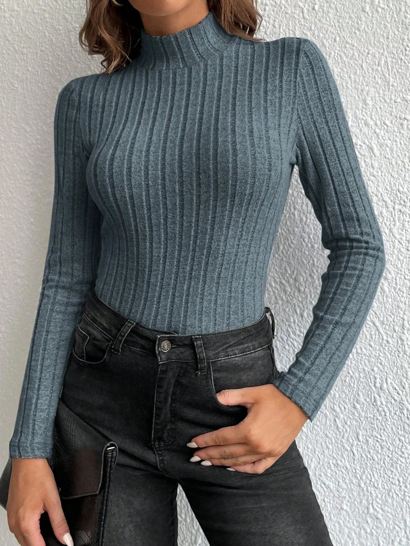 Woman wearing a fitted, long-sleeved, ribbed mock neck top in a blue-grey color with black jeans and a black clutch.