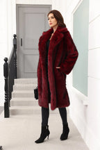 A woman wearing a long, burgundy faux fur coat over a black top and black pants, with hands in pockets.