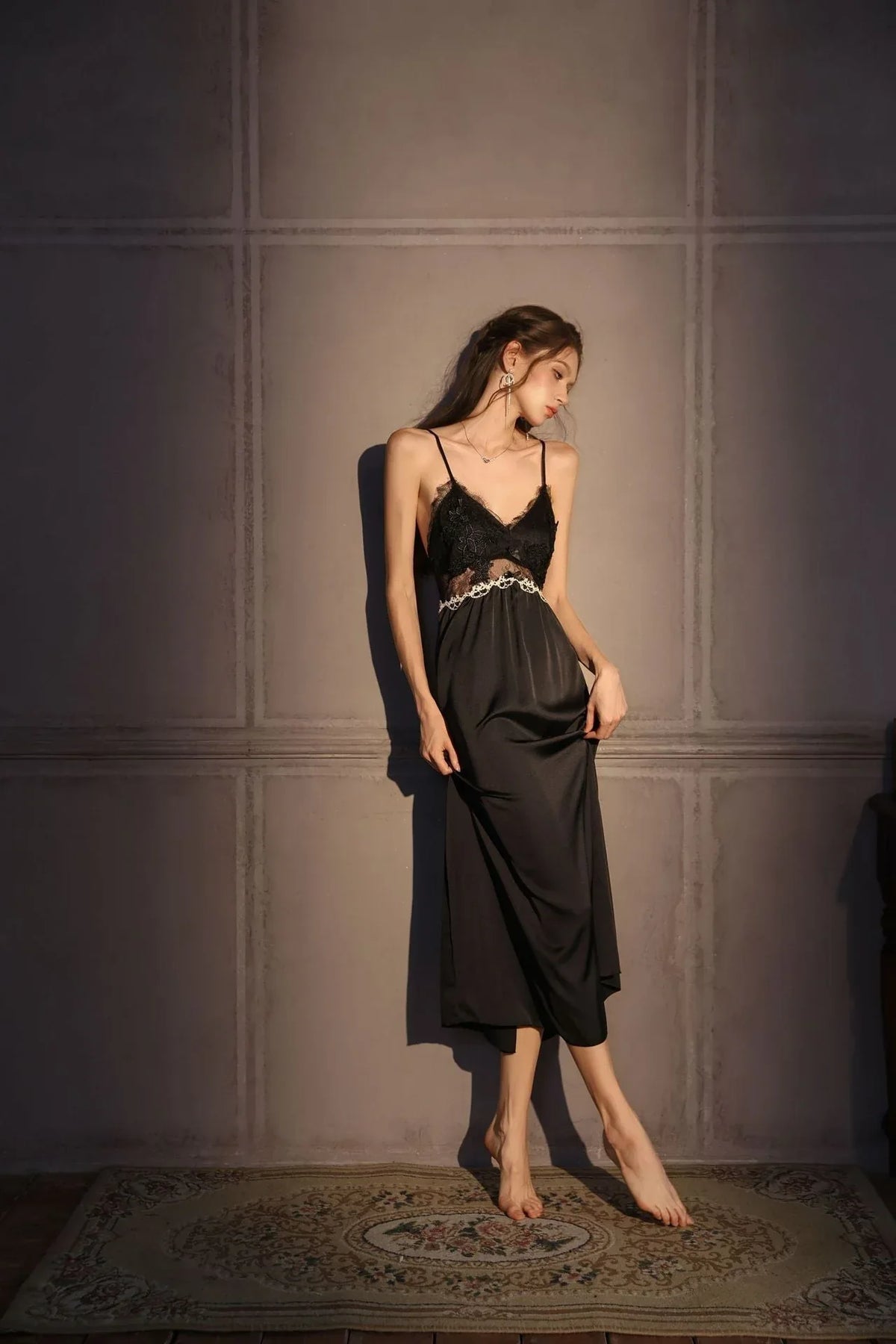 Woman wearing a black satin slip dress with lace trim, standing against a grey wall.