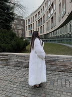 Side profile of a woman wearing a long white faux fur coat, with a modern building in the background.