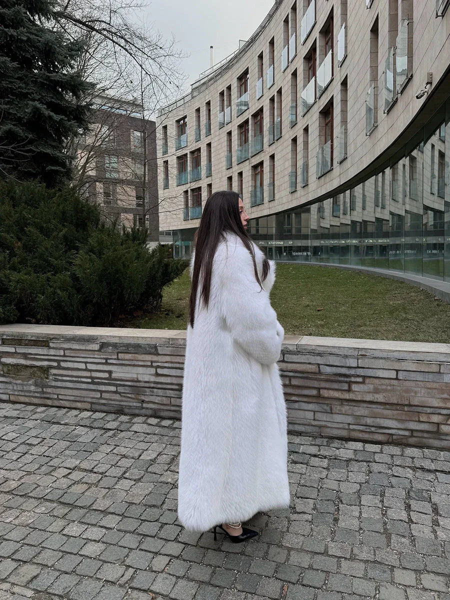 Side profile of a woman wearing a long white faux fur coat, with a modern building in the background.