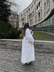 Side profile of a woman wearing a long white faux fur coat, with a modern building in the background.
