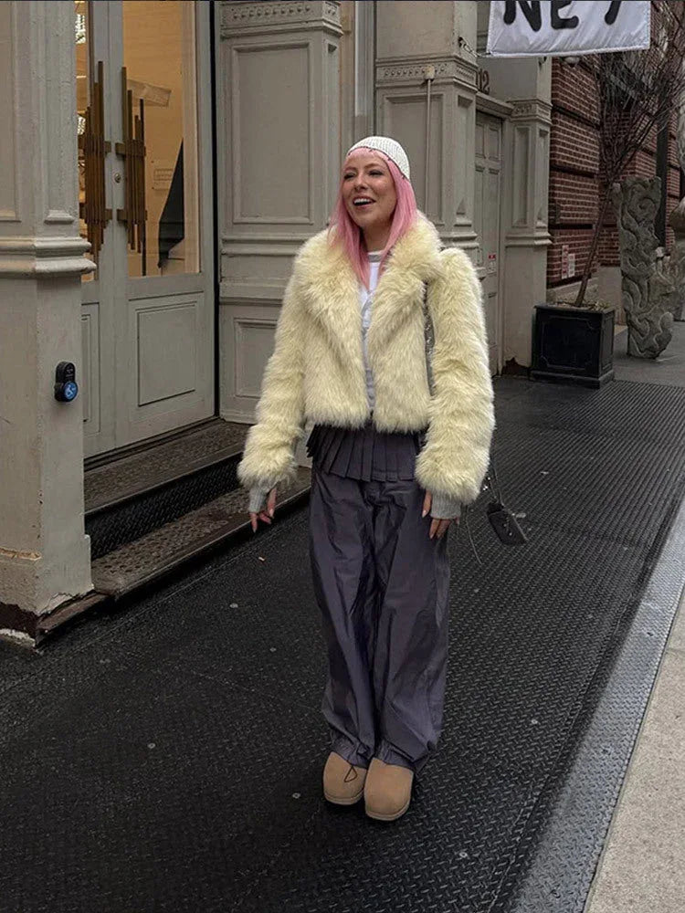 Person with pink hair smiling outdoors, wearing a cream faux fur jacket and grey trousers.