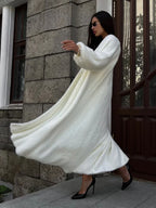 Woman in a long white fuzzy knit dress with flowing fabric, wearing black heels and sunglasses, standing near a wooden door.