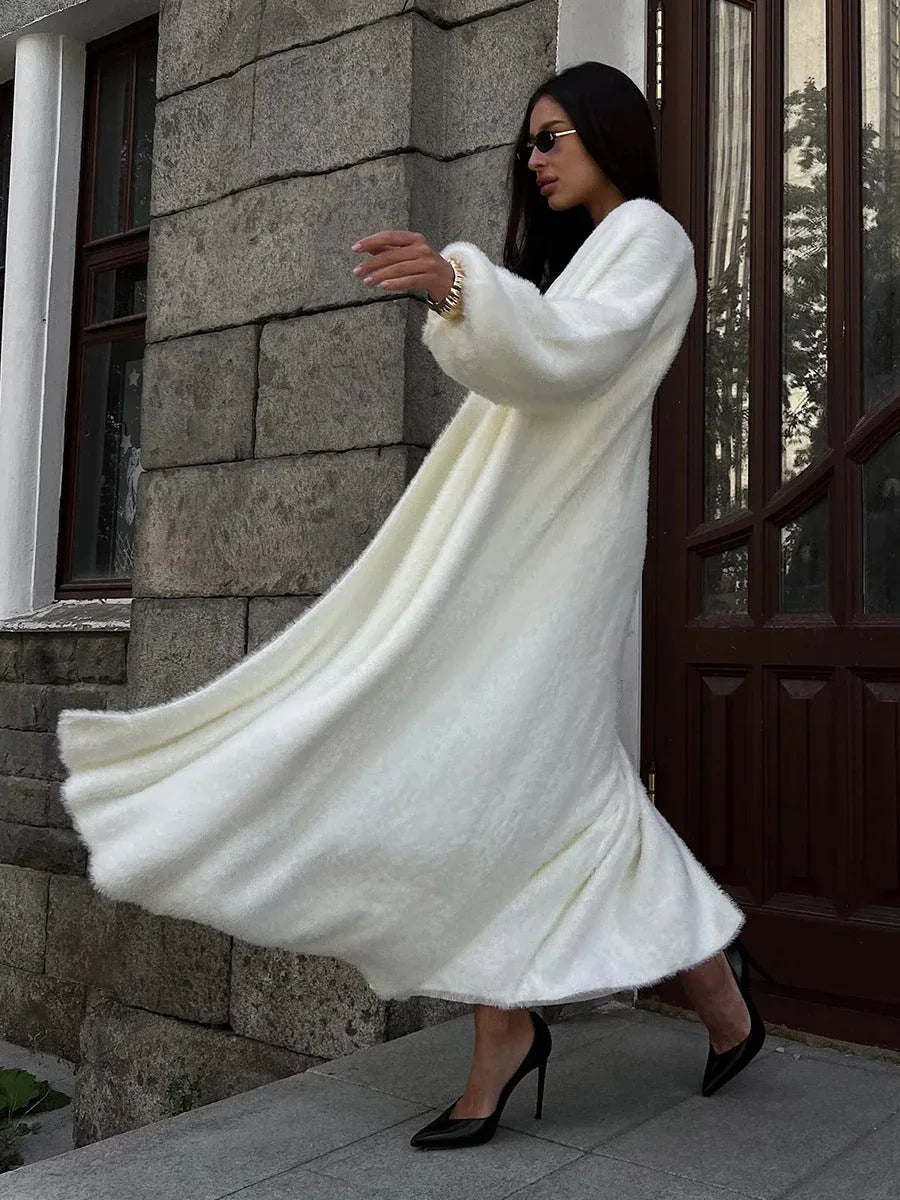Woman in a long white fuzzy knit dress with flowing fabric, wearing black heels and sunglasses, standing near a wooden door.
