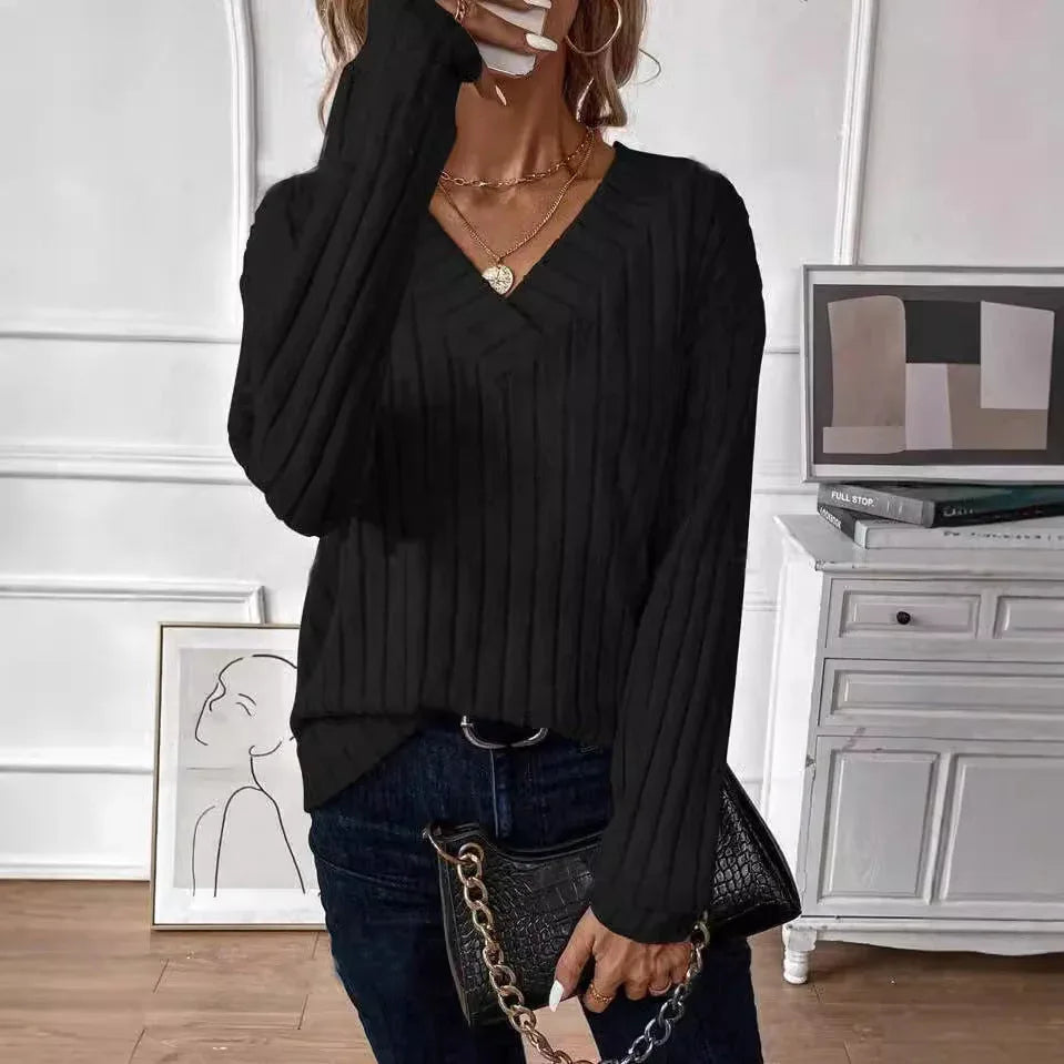 Woman adjusting her black ribbed V-neck long-sleeve sweater, wearing gold necklaces and holding a black handbag.