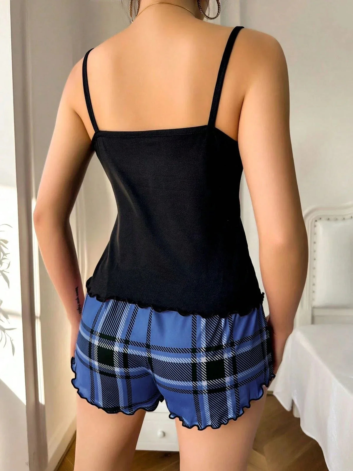 Back view of a woman wearing a black camisole with spaghetti straps and blue plaid pajama shorts with ruffle trim.