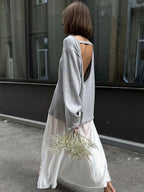 Back view of a woman in a grey oversized knit sweater with an open back detail, wearing a white skirt and holding dried flowers.