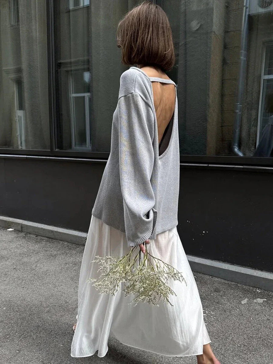 Back view of a woman in a grey oversized knit sweater with an open back detail, wearing a white skirt and holding dried flowers.