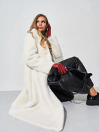 Woman in a white faux fur coat and black pants, wearing red gloves and holding a black bag, kneeling against a white wall.