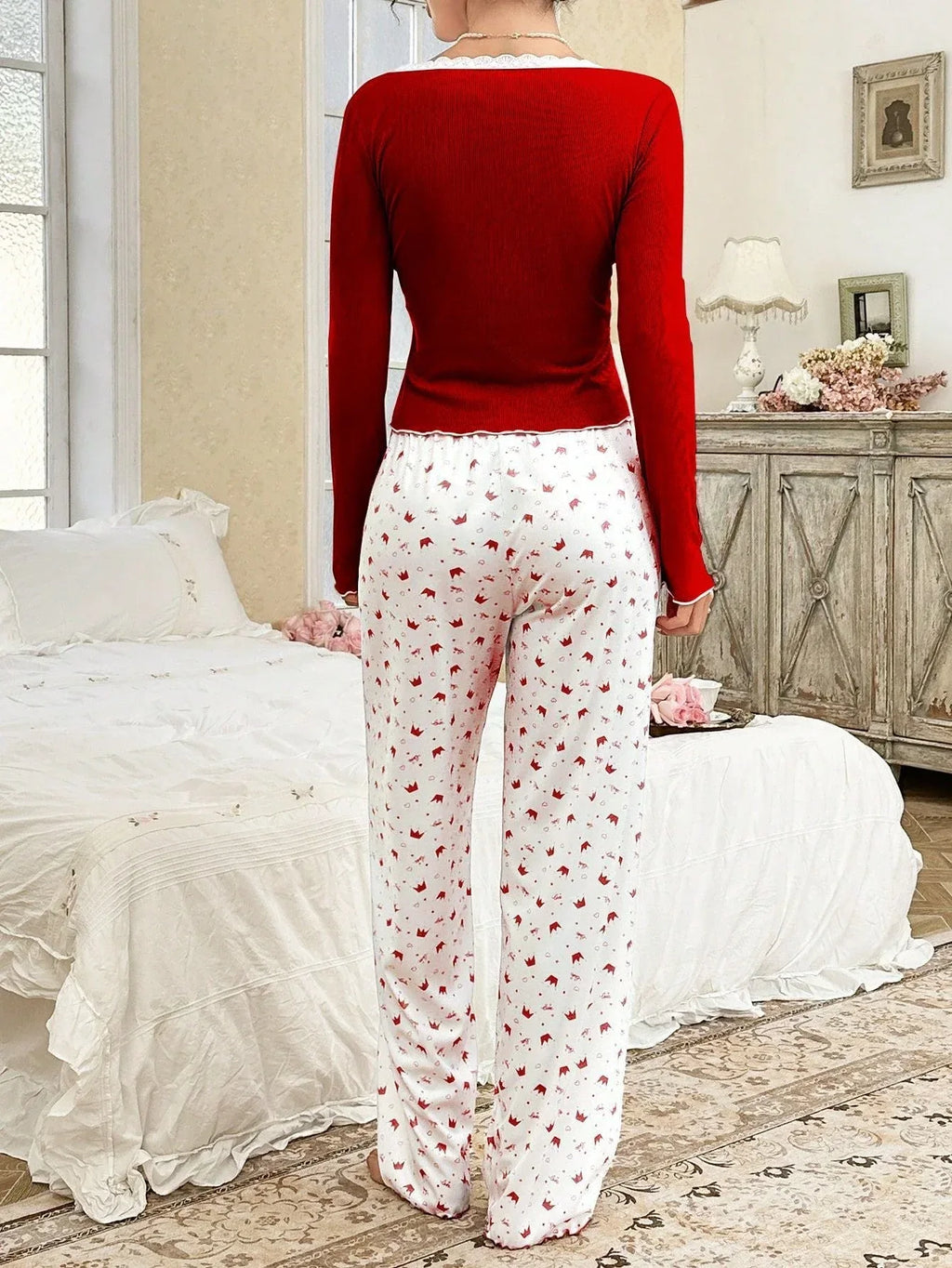 Back view of a woman in a red long-sleeved top and white pants with a red crown print, standing in a bedroom.