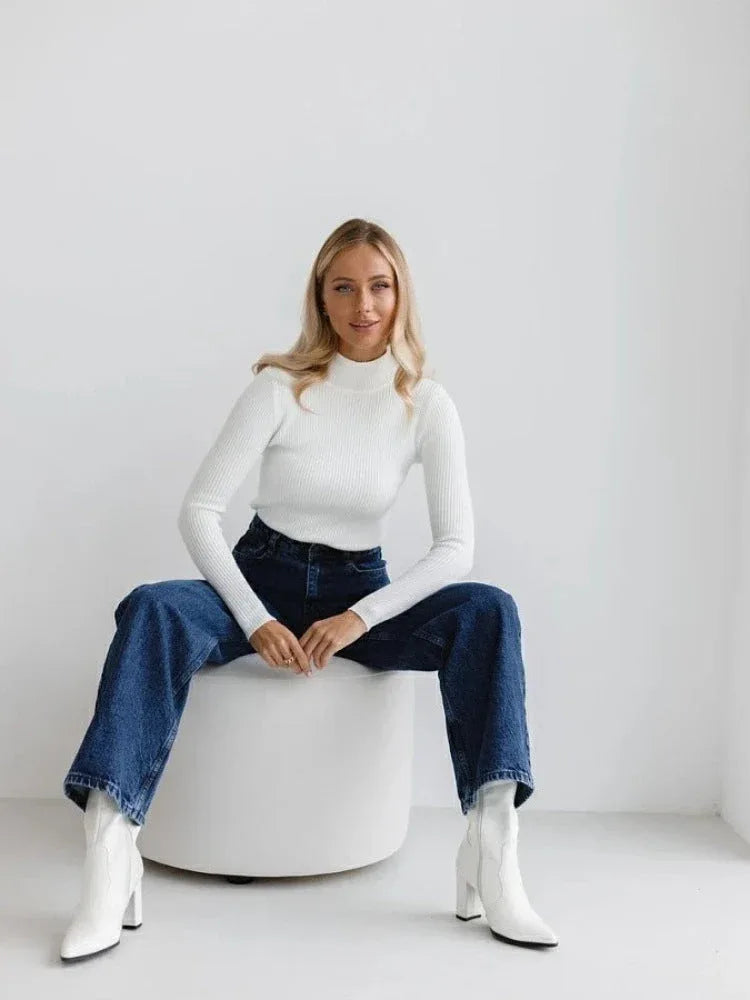 Woman wearing a white ribbed mock neck cropped sweater and dark wash wide-leg jeans, with white ankle boots, sitting on a white ottoman. Secondary image
