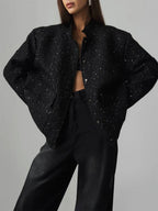 Woman wearing a black sparkly tweed cropped jacket over a black bra and black wide-leg jeans, hands on hips.