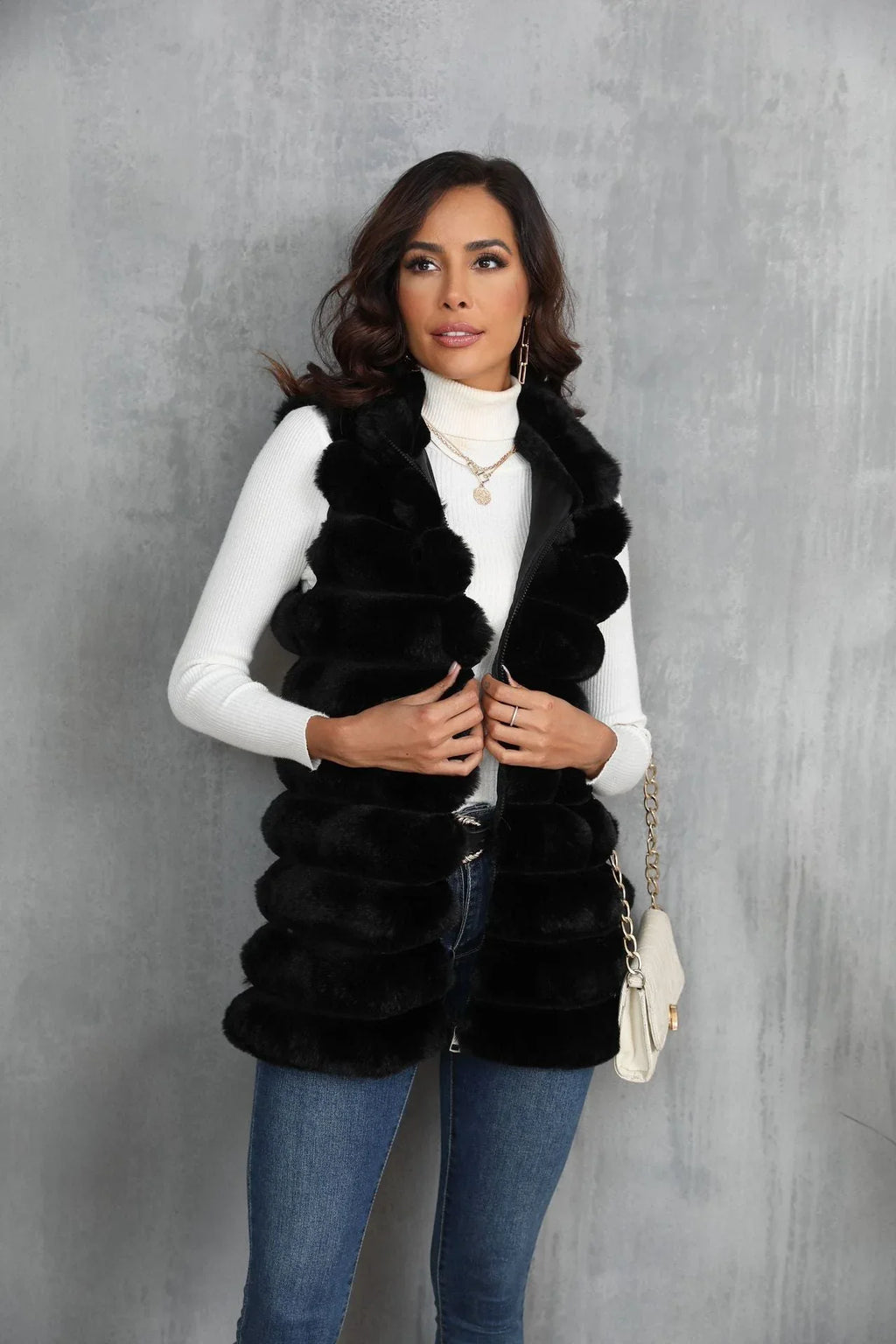 Woman wearing a black faux fur vest over a white turtleneck and blue jeans, holding the zipper.