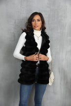Woman wearing a black faux fur vest over a white turtleneck and blue jeans, holding the zipper.