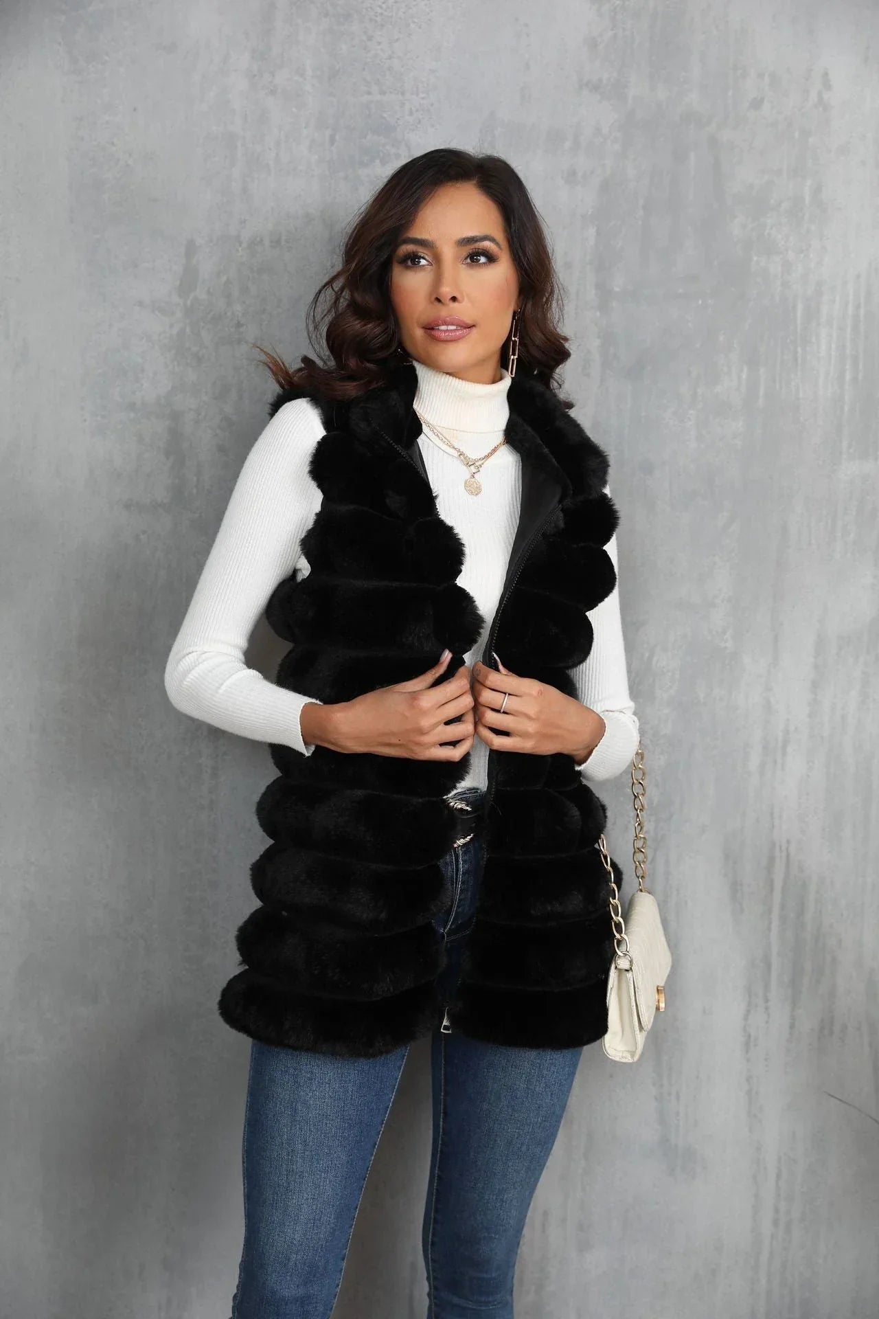 Woman wearing a black faux fur vest over a white turtleneck and blue jeans, holding the zipper.