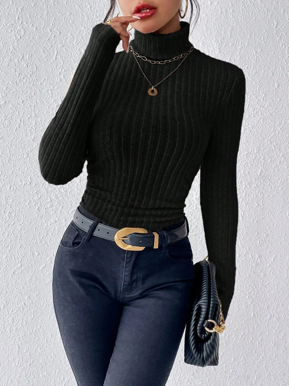 Woman wearing a black ribbed turtleneck sweater, black jeans, a black belt with a gold buckle, and holding a black clutch with a gold chain.
