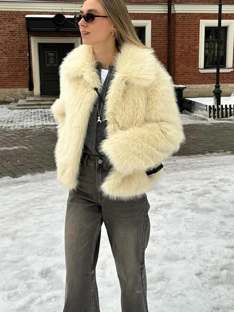 Woman wearing a cream faux fur jacket, grey jeans, and sunglasses, with a necklace visible.