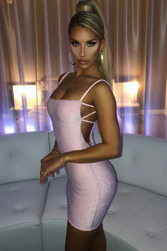 Woman wearing a pink glitter mini dress with spaghetti straps and an open back, standing in a room with plush white seating.