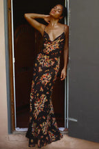 Woman wearing a black floral print maxi slip dress with spaghetti straps and a cowl neck, leaning against a doorway.