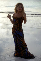 Woman wearing a black, blue, orange, and yellow abstract print maxi dress with spaghetti straps, standing on a beach.