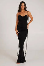 Front view of a woman wearing a long black sleeveless cowl neck dress with adjustable spaghetti straps and tie detail.