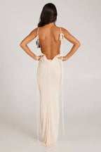 Woman wearing a long cream sleeveless cowl neck dress with an open back and adjustable spaghetti straps.