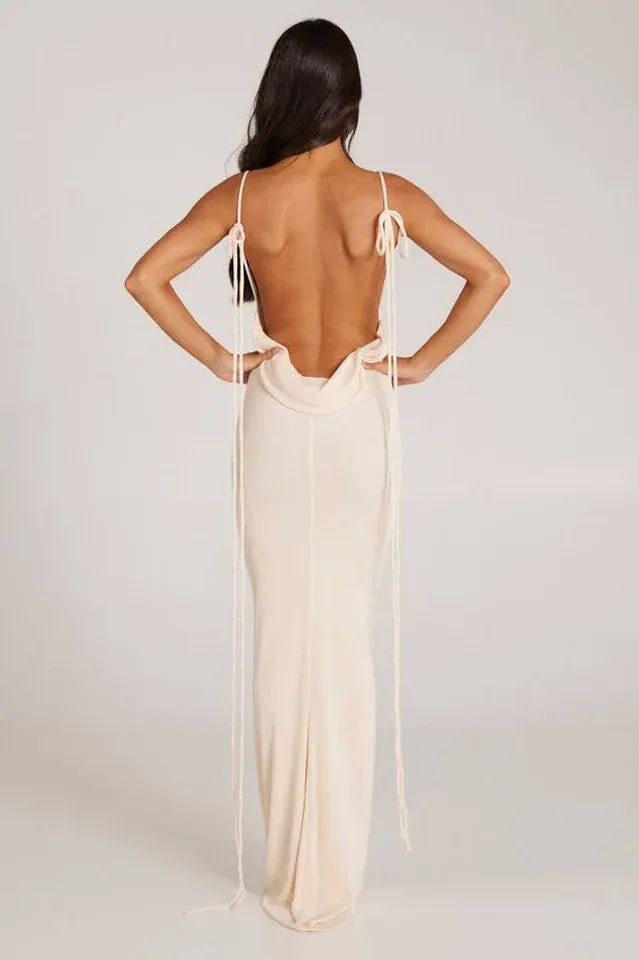 Woman wearing a long cream sleeveless cowl neck dress with an open back and adjustable spaghetti straps.