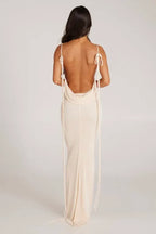 Back view of a woman wearing a long cream sleeveless cowl neck dress with an open back and tie detail.