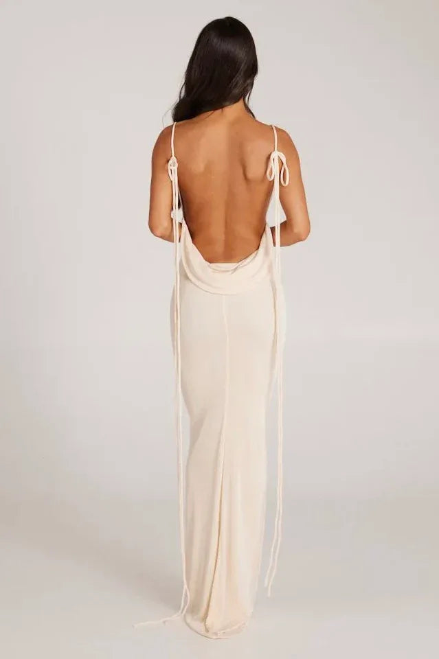 Back view of a woman wearing a long cream sleeveless cowl neck dress with an open back and tie detail.