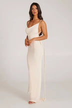 Side view of a woman wearing a long cream sleeveless cowl neck dress with adjustable spaghetti straps and tie detail.