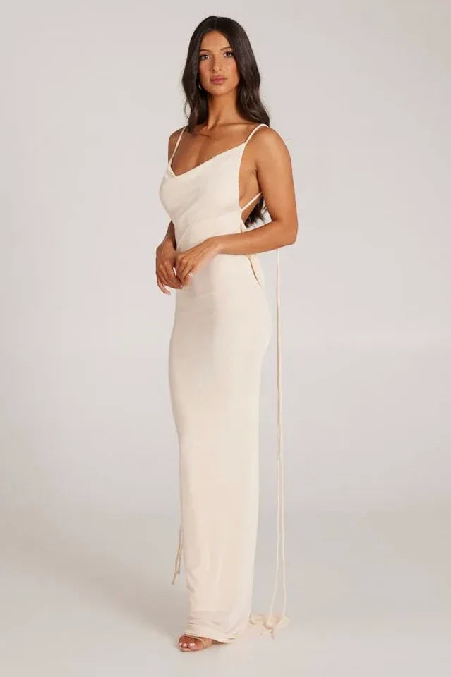 Side view of a woman wearing a long cream sleeveless cowl neck dress with adjustable spaghetti straps and tie detail.