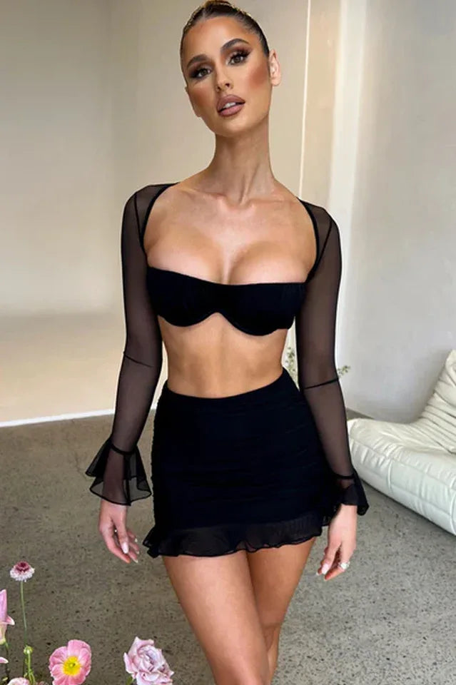 Woman wearing a black mesh two-piece set with a square neck bra top and ruched mini skirt with ruffle hem, in an indoor setting.