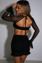 Woman wearing a black mesh two-piece set with a square neck bra top and ruched mini skirt with ruffle hem, from the side.