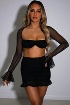 Woman wearing a black mesh two-piece set with a square neck bra top and ruched mini skirt with ruffle hem, showing the back detail.